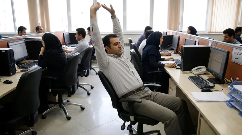 A professional office worker standing at their desk, performing a gentle stretching exercise in a well-lit room.